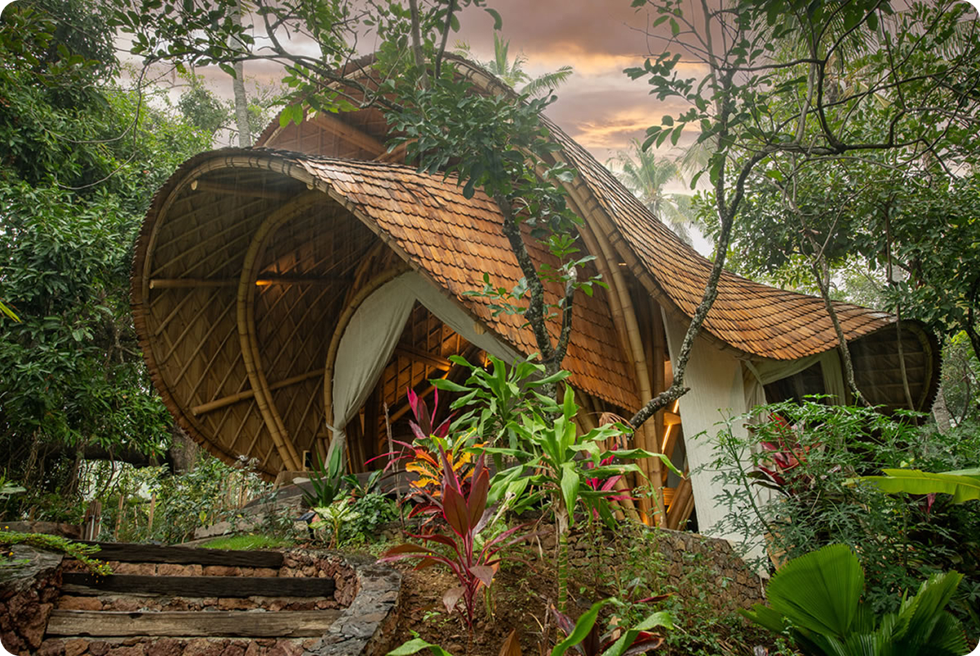 Spiral bamboo bungalow exterior with a shingled roof set among jungle foliage at dusk